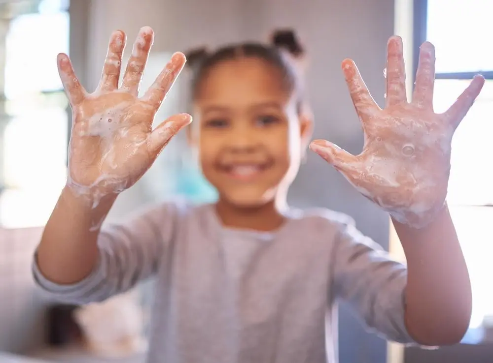 A smiling young girl holds up her soapy hands toward the camera, showing she is washing them. The background is softly blurred, highlighting her face and hands.
